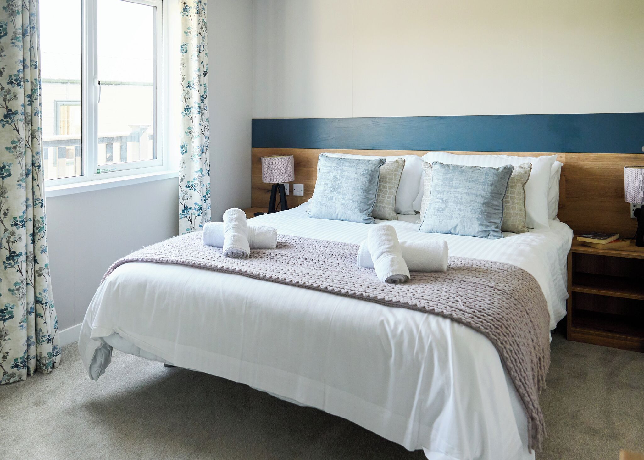 A neatly made bed with white linens and a knitted blanket, a wooden headboard, and a window with floral curtains.