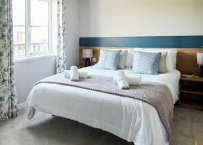 A neatly made bed with white linens and a knitted blanket, a wooden headboard, and a window with floral curtains.