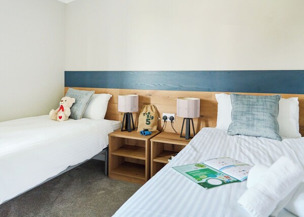 A hotel room with two beds, a nightstand with lamps, a teddy bear, and a bag.