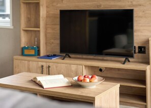 A wooden TV stand with a flat-screen TV, a bowl of fruit, and an open book on a wooden table.
