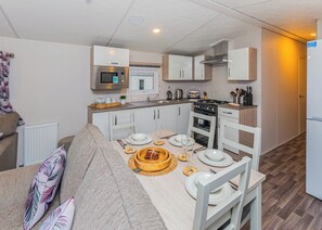 A compact kitchen with a microwave, stove, and sink, a dining table set with plates and cutlery, and a sofa with patterned cushions.