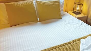 Un lit bien fait, avec du linge de lit blanc et deux grands oreillers, une table de chevet avec une lampe, et une serviette blanche pliée posée sur le lit.