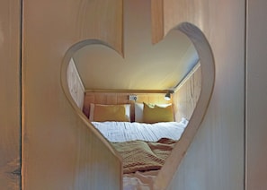 A bedroom with a heart-shaped cutout, wooden walls, a bed with white bedding, and a headboard with a light fixture. - The Woody Safari Tent (Landford, Nr Salisbury)
