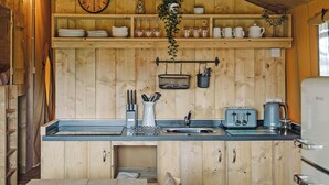 Une cuisine rustique aménagée dans une cabane en bois, avec un évier, une bouilloire, un grille-pain et des étagères remplies de vaisselle et d’une horloge.