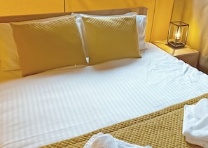 Ein ordentlich bezogenes Bett mit weißen Leinen und zwei großen Kissen, ein Nachttisch mit Lampe und ein gefaltetes weißes Handtuch auf dem Bett.