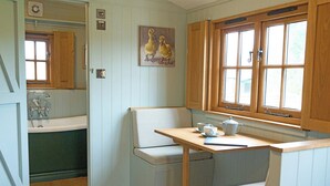 Een keuken met houten kastjes, een raam, een schilderij van eenden en een tafel met een waterkoker en kopjes.