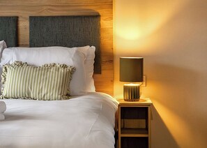 Ein ordentlich bezogenes Bett mit weißen Leinen und gestreiften Kissen, einer Nachttischlampe und einem hölzernen Kopfteil.
