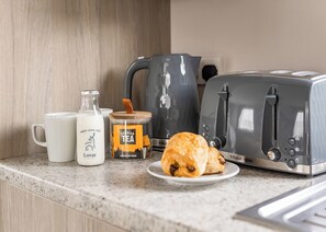 Un plan de travail de cuisine avec un grille-pain, une machine à café, une bouteille de lait, un pot de thé et un croissant sur une assiette.