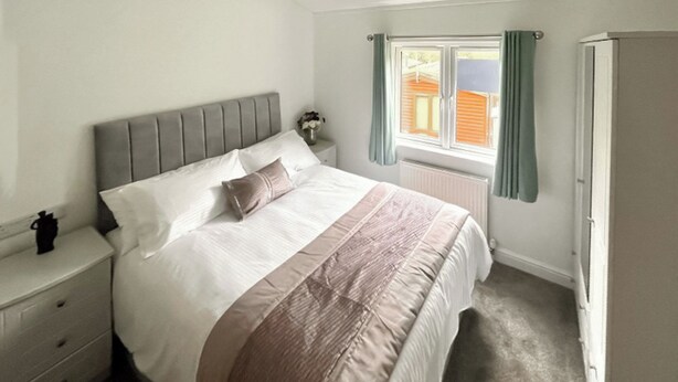 Une chambre à coucher comprenant un lit, une table de chevet, une commode, une fenêtre avec des rideaux et un radiateur.