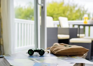 Ein Tisch mit Feldsteuerre, einer braunen Tasche und einer Zeitung, im Hintergrund ist ein Balkon mit Stühlen zu sehen.