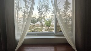 A room with a window featuring sheer white curtains, a wooden floor, and a view of a park with trees and a pathway.