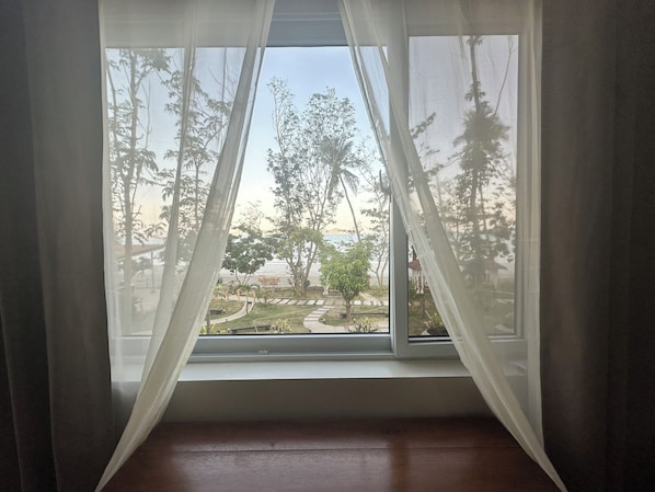 Una habitación con ventana con cortinas blancas translúcidas, piso de madera y vista a un parque con árboles y un sendero.