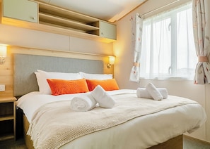 Un lit bien fait, agrémenté d’oreillers blancs et orange, une serviette blanche roulée et une couvre-lit beige. On y trouve une tête de lit en bois, une table de chevet avec une lampe, et une fenêtre avec des rideaux.