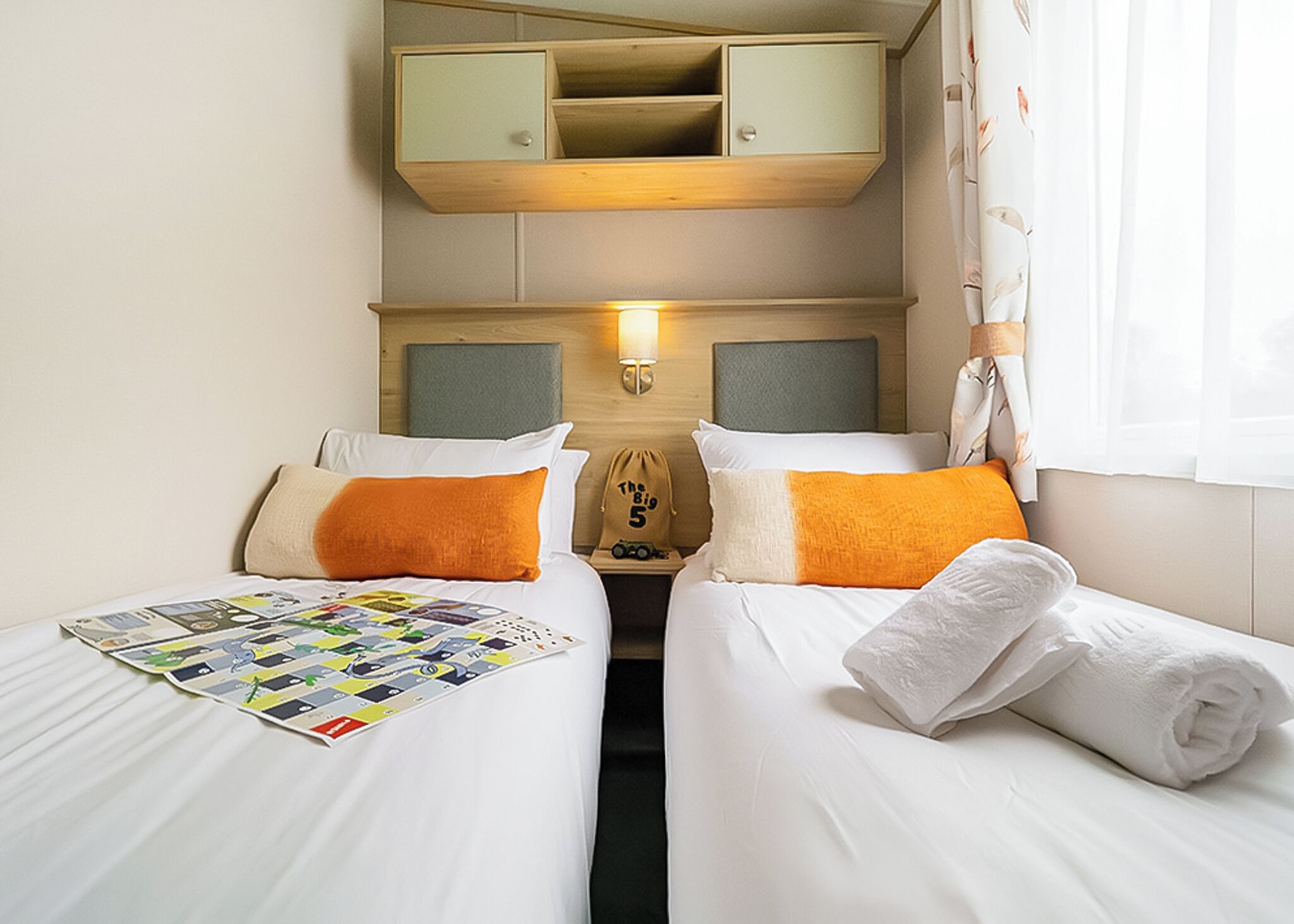 A hotel room with two beds, a small table with a board game, a nightstand with a lamp, and a wardrobe above the beds.