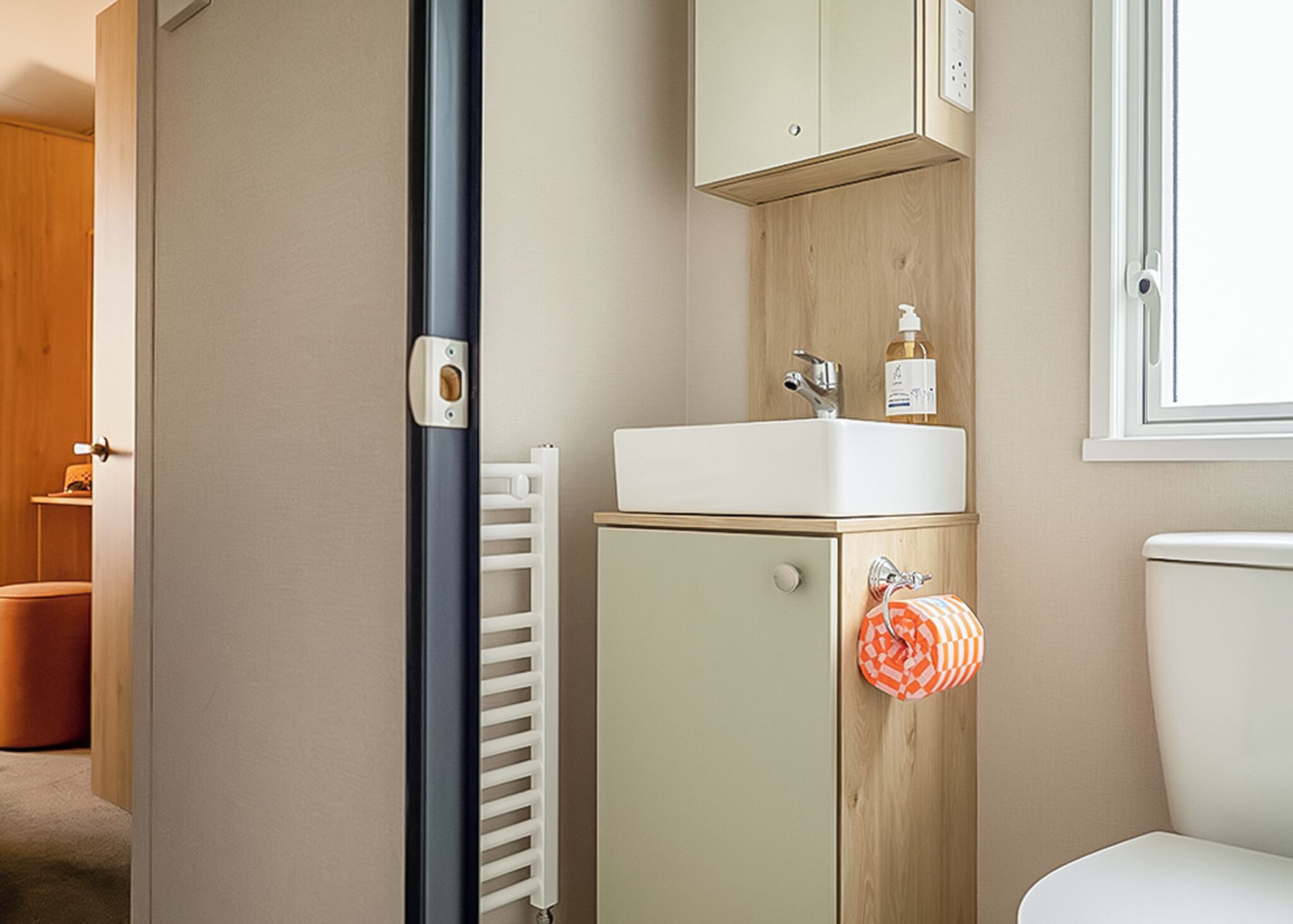 Un baño con lavamanos blanco, mueble de madera y un perchero con una toalla de cuadros.