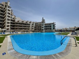 Una piscina rodeada de un área al borde con sillas de descanso, un solárium y un edificio de varios pisos al fondo.