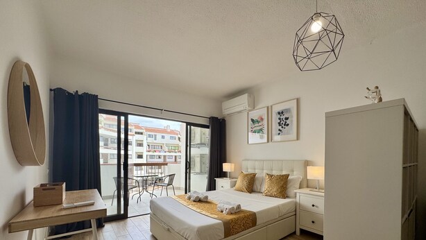 A bedroom with a bed, bedside tables, a desk, a chair, a wardrobe, and a balcony with a view of buildings. - Albufeira Skylight 3 With Pool by Homing (Albufeira)
