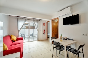 A room with a red sofa, a white table with four chairs, a flat-screen TV mounted on the wall, and a sliding glass door leading to an outdoor area. - Vacation Home 'My Little Bungalow 50' with Mountain View, Wi-Fi, and Air Conditioning (Palm-Mar)