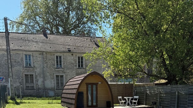 A small, wooden, semi-circular structure with a porch and seating area, set in a grassy yard with a stone building in the background. - Quiet cabin in the Fontainebleau forest (Oncy-sur-École)