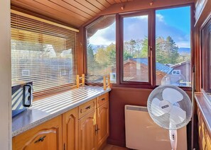 A kitchen with wooden cabinets, a countertop, a window with blinds, a fan, and a view of houses and trees.