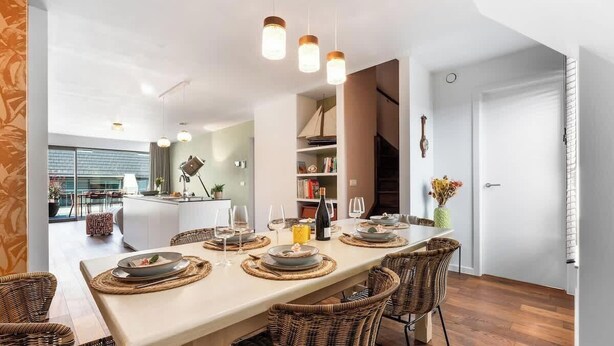 Un espace repas moderne avec une longue table dressée pour quatre personnes, entourée de chaises en osier, et une cuisine visible au fond.