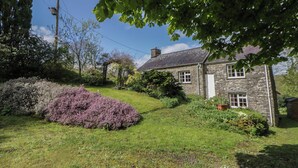 A stone building with a grassy yard, a purple bush, and a clear sky. - Tyn Y Fron (Llanon)