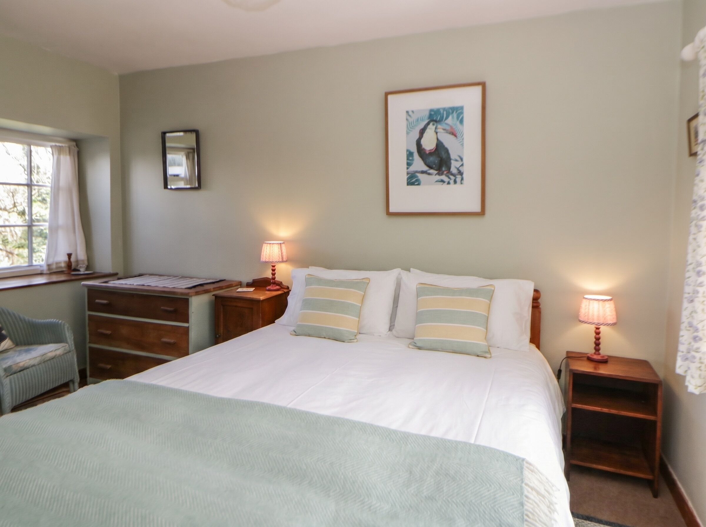 Une chambre à coucher avec un lit, deux tables de chevet avec des lampes, une chaise, un bureau, un tableau encadré au mur et une fenêtre avec des rideaux.