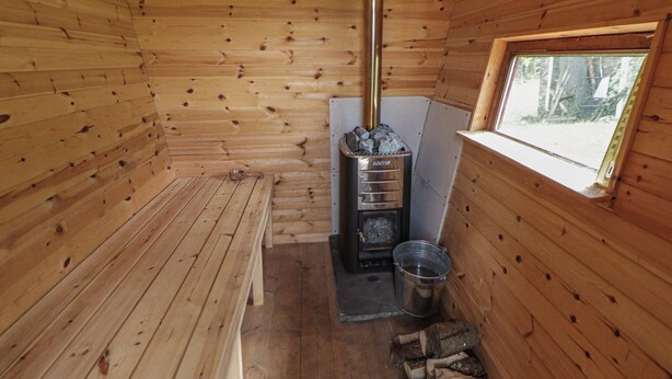 A wooden cabin interior with a wood-burning stove, a window, and a metal bucket. - Tyn Y Fron (Llanon)