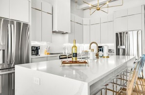 A modern kitchen with stainless steel appliances, a marble countertop, and a central island with a wine bottle and glasses. - BRAND NEW, Elevated Pool, Beach Front, Hot Tub, and Elevator - Dune Alright (St. George Island)