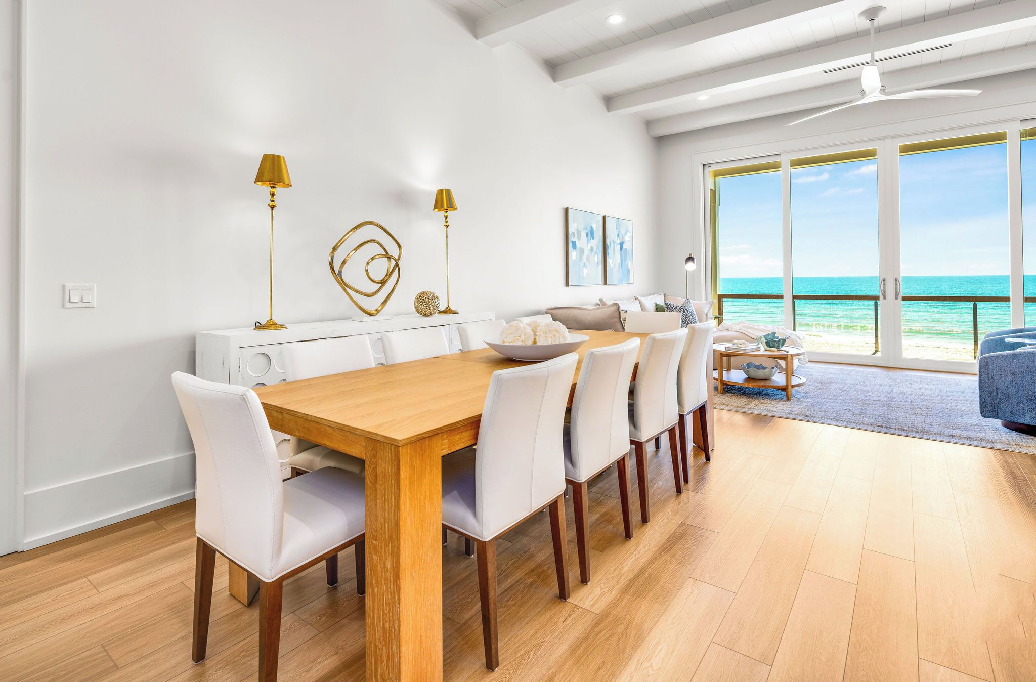 A modern dining area with a wooden table and white chairs, a white console table with gold lamps, and a view of the ocean through large glass doors.