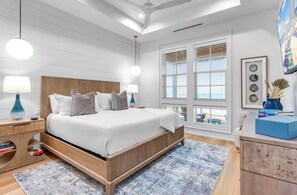 A modern bedroom with a wooden bed, two bedside tables with lamps, a large window with blinds, and a view of the outside. - BRAND NEW, Elevated Pool, Beach Front, Hot Tub, and Elevator - Dune Alright (St. George Island)