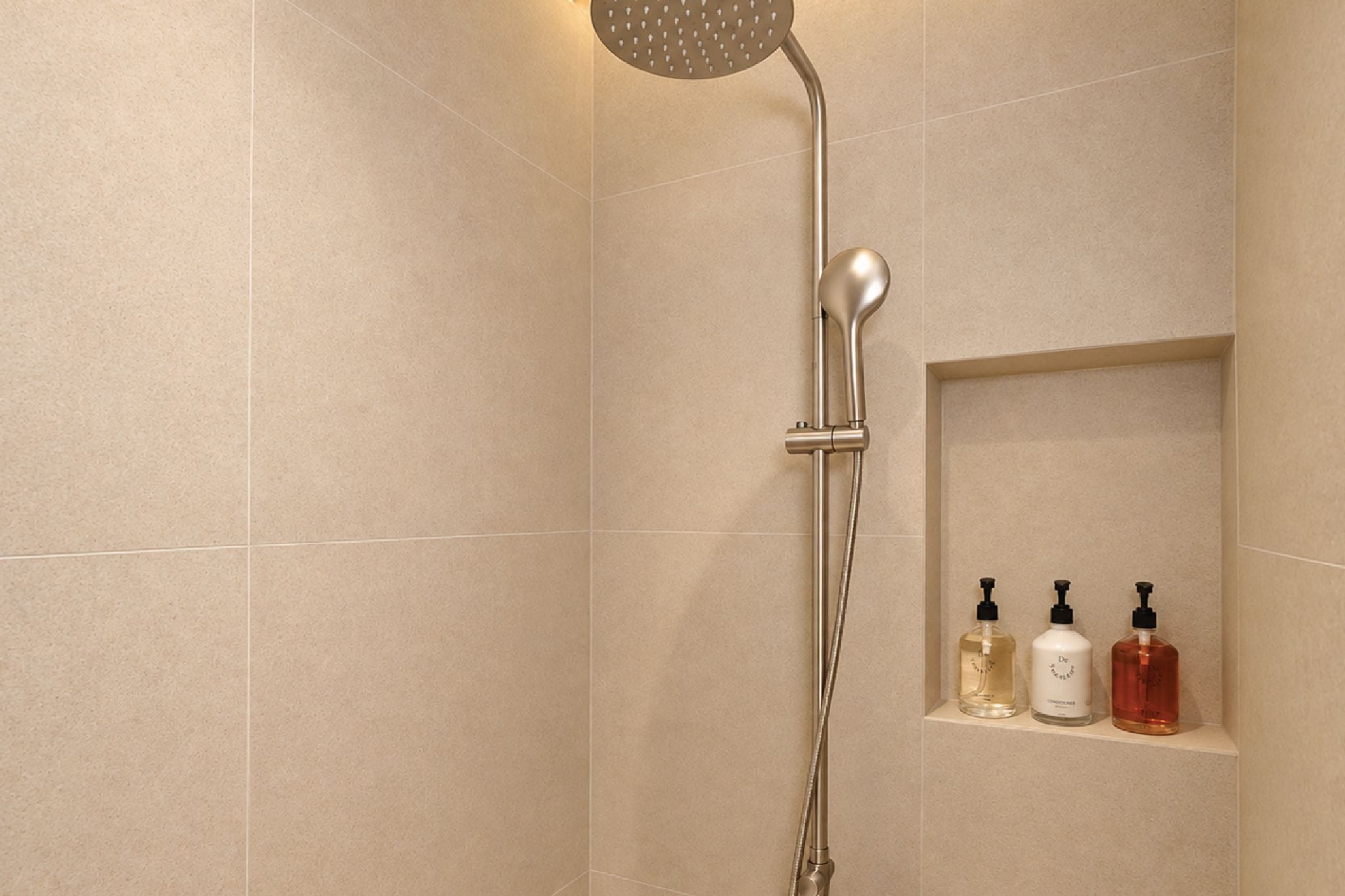 A shower with a handheld showerhead and a wall-mounted control. There are three bottles of liquid soap on a shelf.