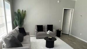 Una sala de estar con un sofá gris, una mesa de centro negra y un alfombra blanco de pelo alto.