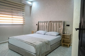 A bedroom with a bed, a nightstand, a wooden shelf, and a window with blinds. - 2-bedroom apartment in gorgeous Marrakech with AC and charm (Marrakech)