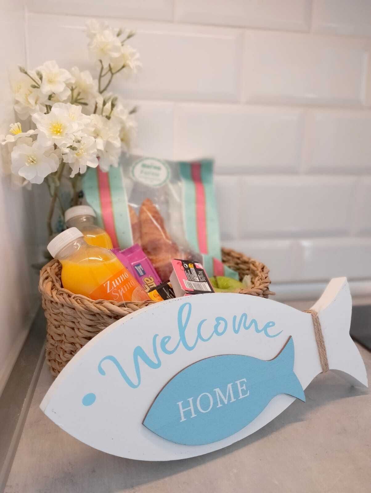 Un panneau décoratif en forme de poisson, sur lequel est inscrit le mot « MAISON », est posé sur un comptoir de cuisine, entouré d’un panier d’épicerie et d’un bouquet de fleurs.