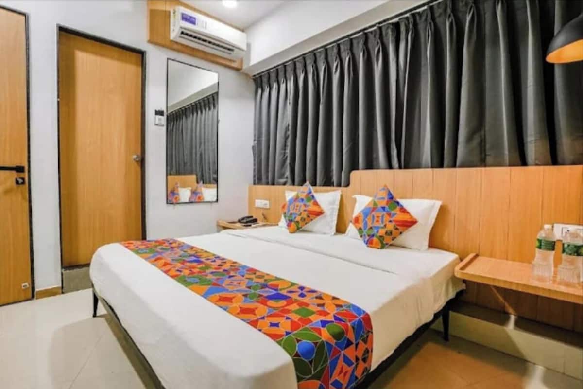 A hotel room with a bed, a bedside table, a mirror, a window with curtains, and an air conditioning unit.