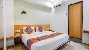 A hotel room with a bed, a bedside table with a lamp, an air conditioning unit, and a wooden door.