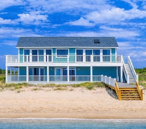 Ein Strandhaus mit zwei Etagen, einem Balkon, einer Holzterrasse und einer Treppe, die zum oberen Stock führt.