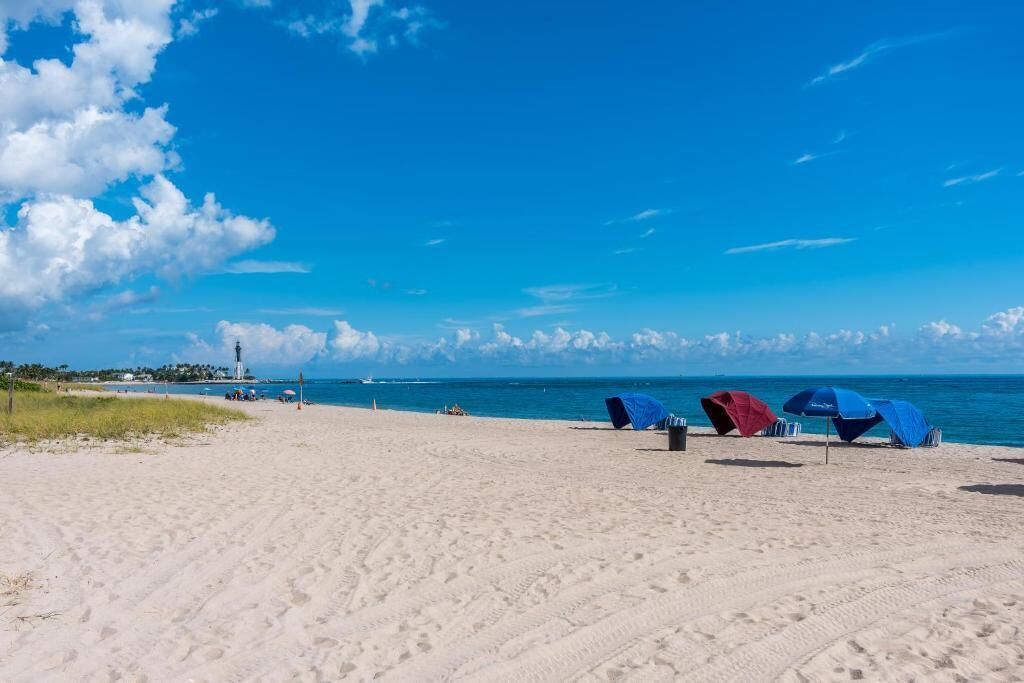 A beach with sand, a lighthouse, and blue umbrellas.