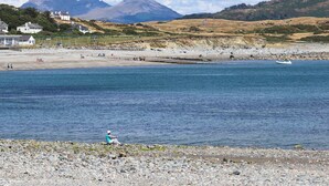 A coastal landscape with a rocky beach, a person sitting on the shore, and houses in the background. - Ty Gwenno (Criccieth)
