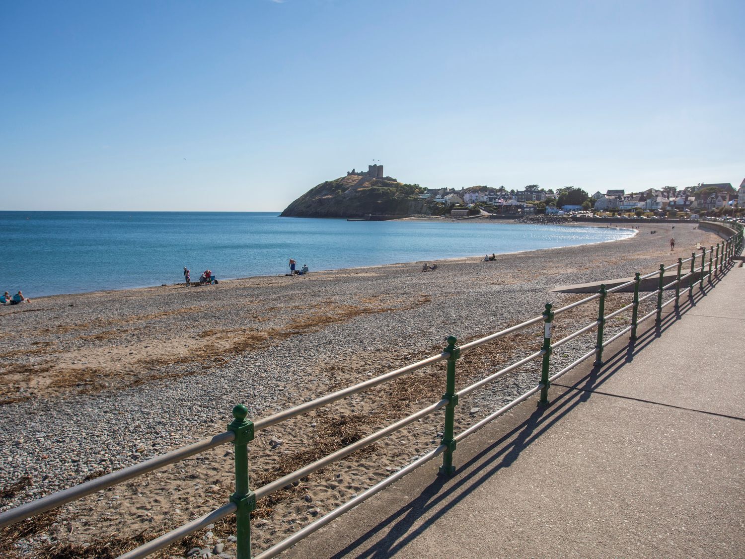 A coastal beach with a promenade, pebbles, and a distant hill with a structure.