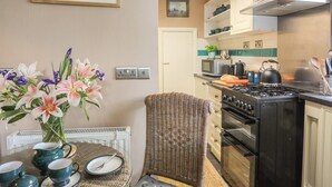 A kitchen with a stove, microwave, and a dining area with a wicker chair and a vase of flowers. - Ty Gwenno (Criccieth)