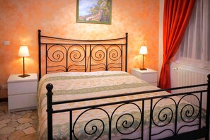 Ein Bett mit Metallgestell, zwei Nachttische mit Lampen, eine gemusterte Bettdecke, ein Bild an der Wand und ein Fenster mit Vorhängen.
