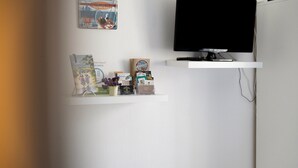A room with a television on a white stand, a shelf with various items including books and a small plant, and a wall-mounted picture. - Guest Room “Chambre Saint-Samson-sur-Rance” with Private Terrace, Garden & Wi-Fi (Saint-Samson-sur-Rance)