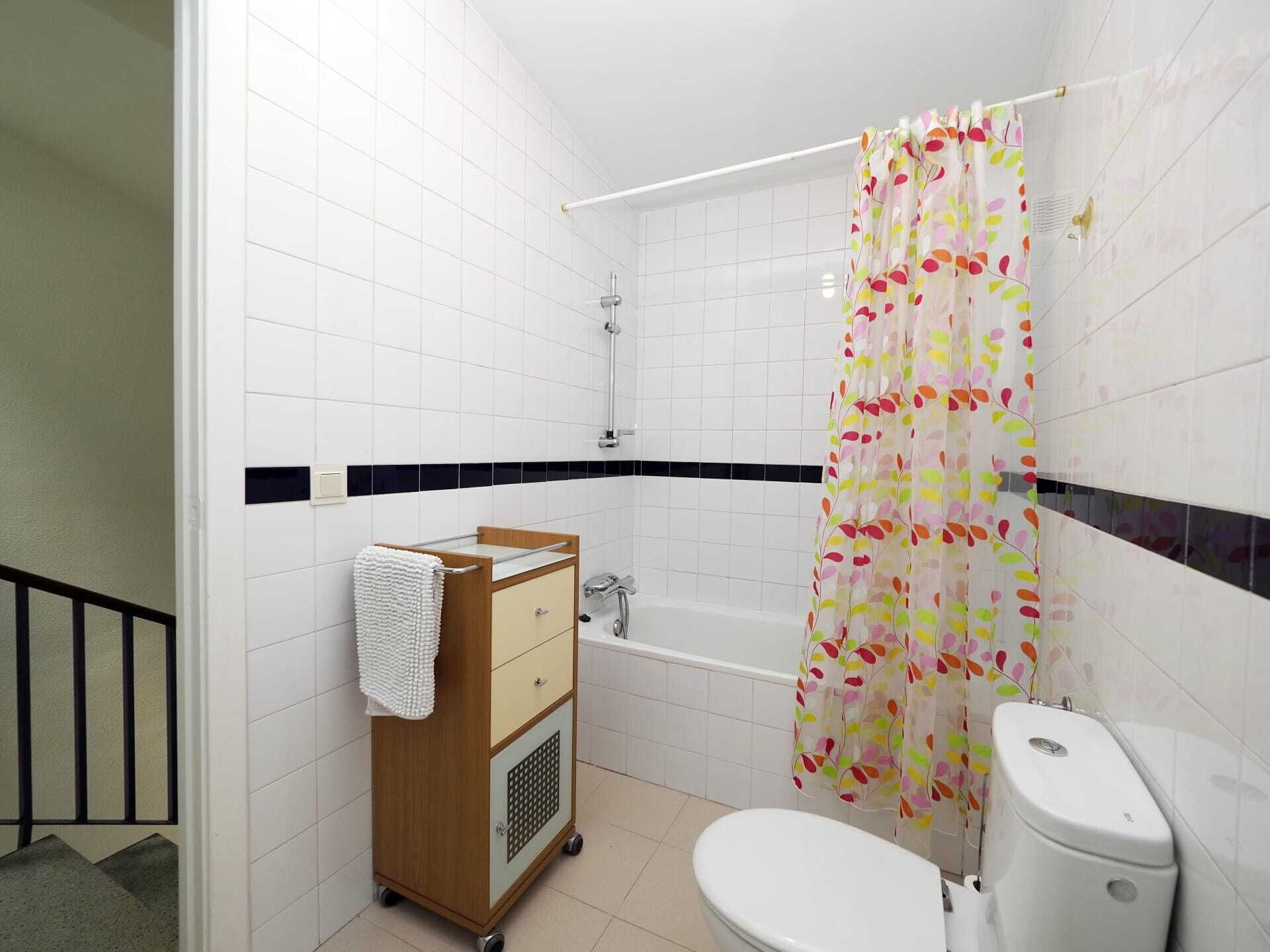 A bathroom with white tiles, a bathtub, a toilet, and a wooden cabinet with a towel.