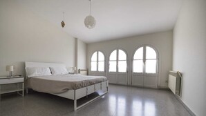 Un dormitorio con una cama, una mesita de noche, una lámpara, un radiador y ventanas de arco.