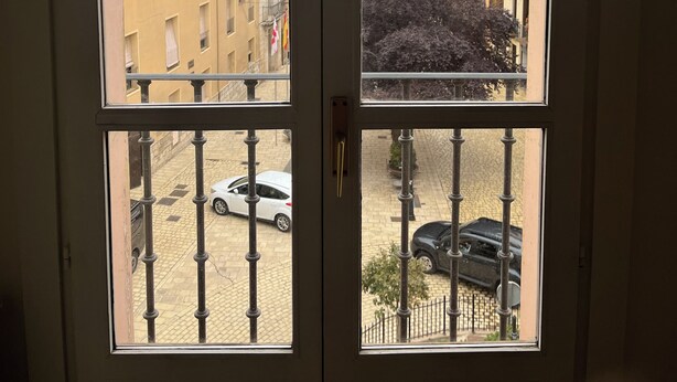 Vista a través de un ventanal de doble puerta con barandilla metálica, que da a una calle con coches estacionados y edificios.