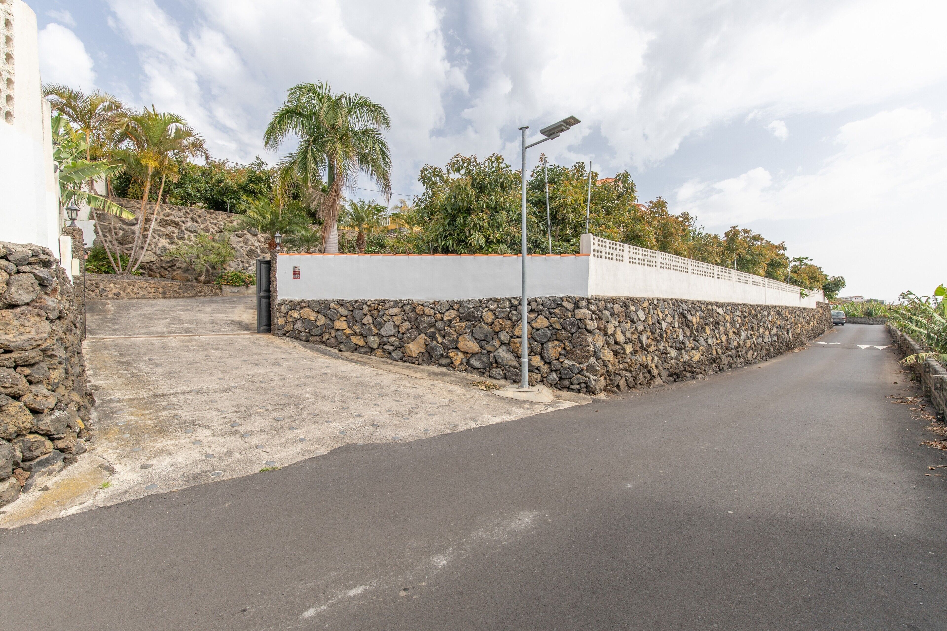 Una strada asfaltata con un muro in pietra, un edificio bianco e palme.