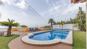 En pool med buet kant, omgivet af en flisepavimenteret terrasse, og udsigt til havet.