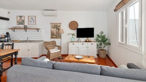 Una cocina moderna con armarios blancos, una mesa de comedor y un sofá en una sala de estar con televisor y plantas.